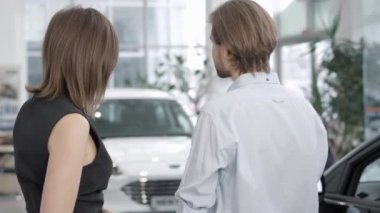 Bayilikte ya da galeride konuşan neşeli beyaz kadın ve erkeğin arka plandaki görüntüsü. Olumlu genç alıcı ve satıcı içeride araç alımını tartışıyor. Otomobil endüstrisi.