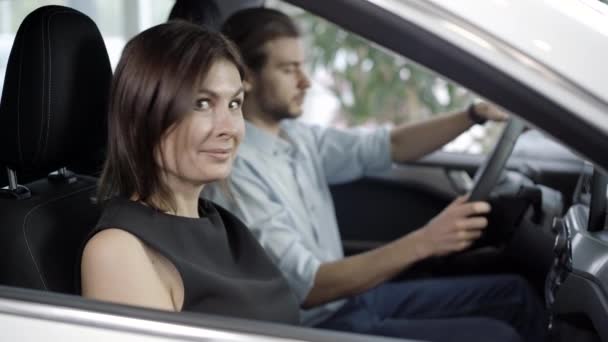 Retrato de una mujer alegre sentada en el salón de coches, girando hacia la cámara y mostrando los pulgares hacia arriba. Hombre caucásico borroso en el asiento del conductor en el fondo. Pareja comprando vehículo en concesionario o sala de exposición. — Vídeo de stock