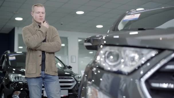 Portret van bedachtzame jonge blanke man kiezen auto in de dealer of showroom. Ernstig vertrouwen mannelijke koper staat binnen en kijkt naar zwart voertuig. — Stockvideo