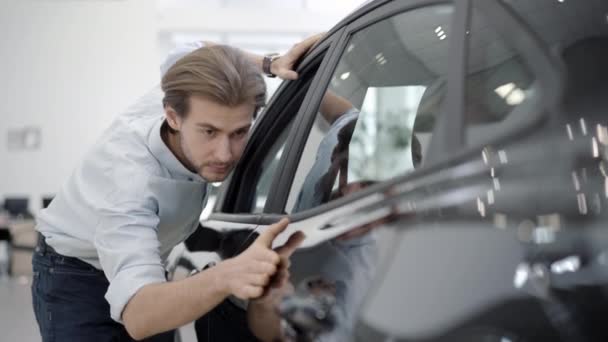 Portrét pohledného muže dotýkajícího se nového černého auta v showroomu. Sebevědomý mladý bělošský kupec obdivující automobil v prodejně aut. Životní styl a koncept úspěchu. — Stock video