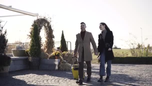 Couple caucasien riche avec différence d'âge marchant avec sac de voyage jaune à l'extérieur le jour ensoleillé d'automne ou de printemps. Portrait de riche confiant homme et femme tenant la main et regardant la maison de vacances. — Video