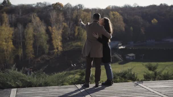 Ampla vista de volta tiro de feliz abraço casal caucasiano em pé na ponte de madeira no campo falando. Homem e mulher relaxados admirando a beleza da natureza olhando para a floresta de outono ao fundo. — Vídeo de Stock