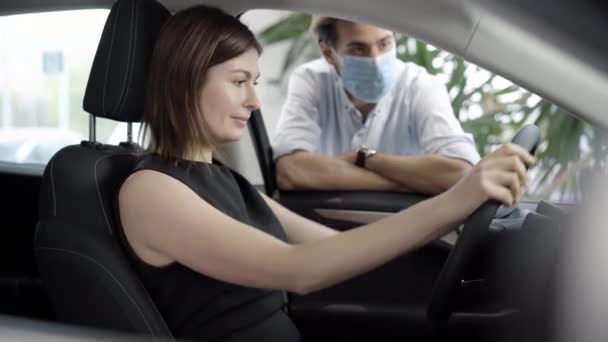年轻而兴奋的女人坐在司机的座位上，就像一个戴着科维德面具说话的男人。Coronavirus病毒大流行期间在汽车经销店购买汽车的成功女商人的画像. — 图库视频影像