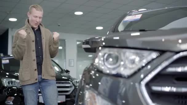 Feliz joven caucásico hombre mirando un coche nuevo en la concesionaria y mostrando el gesto de victoria. Satisfecho cliente masculino exitoso compra de vehículo en sala de exposición. Alegría y estilo de vida. — Vídeos de Stock