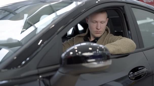 Portret van succesvolle mannelijke koper zittend in nieuwe zwarte auto en onderzoekend voertuig. Jonge blanke man kiezen auto in de auto showroom of dealer. Auto-industrie en bedrijfsconcept. — Stockvideo