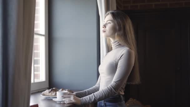 Side view of thoughtful confident young woman looking out the window in the morning. Portrait of gorgeous beautiful brunette Caucasian lady standing indoors with coffee cup and croissant on windowsill — Stock Video