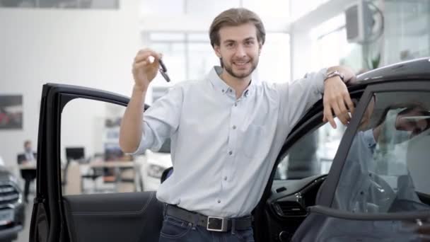 Retrato de hombre feliz jactándose de las llaves del coche de pie en el nuevo automóvil en la concesionaria. Comprador caucásico satisfecho posando con vehículo negro en la sala de exposición. Concepto de éxito y riqueza. — Vídeo de stock