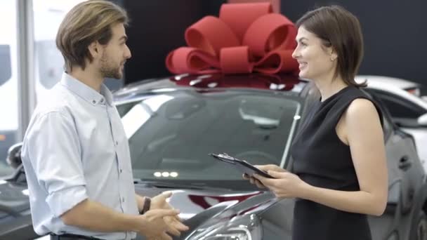 Glada rika man pratar med bilhandlare i showroom. Sidovy av positiv vit köpare och ung brunett professionell kvinna diskuterar köp av svart fordon i återförsäljaravtal. — Stockvideo