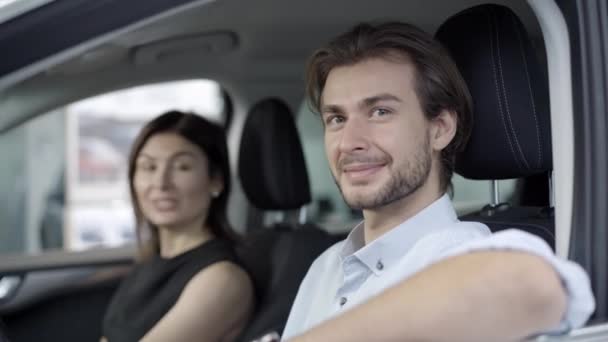 Retrato de un joven hombre guapo sonriente sentado en el asiento del conductor y mirando a la cámara con la hermosa mujer caucásica borrosa en el fondo. Feliz propietario del vehículo nuevo en la concesionaria de automóviles. — Vídeo de stock