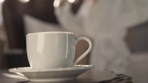 Primer plano de la taza de café blanco en la mesita de noche con una joven delgada borrosa que se extiende al fondo y se va. Irreconocible dama caucásica despertando en la mañana en casa o en la habitación de hotel. — Vídeos de Stock