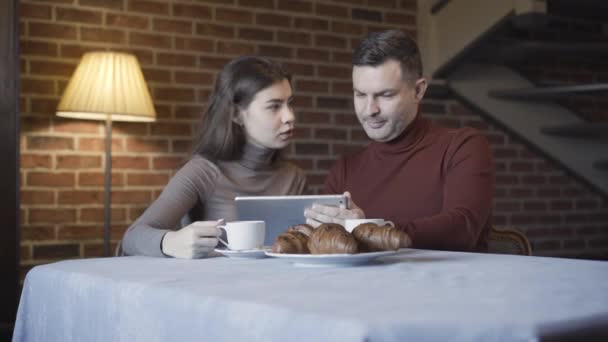 Bogata kaukaska para z różnicą wieku surfuje po Internecie na tablecie i rozmawia podczas śniadania w domu. Pewny siebie przystojny dorosły mężczyzna i młoda piękna kobieta podczas lunchu w pomieszczeniach. — Wideo stockowe