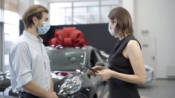 Zelfverzekerde blanke verkoper en koper in Covid-19 gezicht maskers praten in de auto-dealer. Zijaanzicht van succesvolle zakenman en jonge dealer bespreken van nieuwe zwarte auto staan op de achtergrond. — Stockvideo