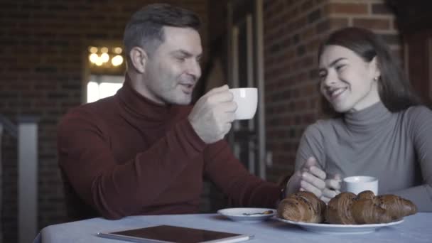 幸せな白人のカップルは朝コーヒーを飲んで話しています。ハンサムな大人の男性と若い美しい女性の肖像笑顔と朝食に自宅でチャット。団結と家族の概念. — ストック動画