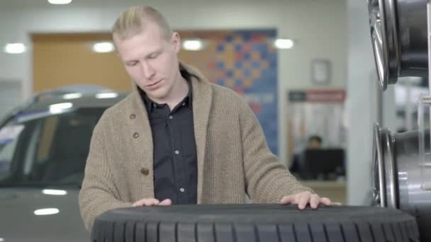 중심적 인 젊은 백인 남성이 쇼룸이나 대리점에서 자동차 타이어를 검사하고 있습니다. 집중 고객이 실내에서 자동차 부품을 선택하는 모습. 자동차 산업 개념. — 비디오