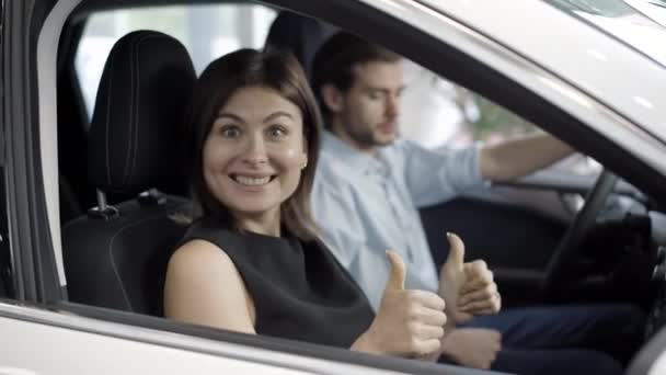 Opgewonden mooie vrouw glimlachend naar de camera en met duimen omhoog wijzend naar de man zittend op de bestuurdersstoel. Gelukkig Kaukasische vrouw het kopen van nieuwe auto met man in auto-dealer. — Stockvideo