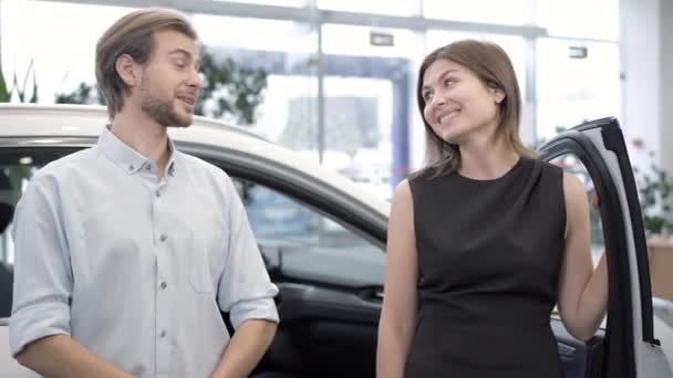 Un cuplu tânăr fericit stând la o mașină nouă în reprezentanță vorbind, uitându-se la cameră și zâmbind. Portretul soțului și soției caucaziene de succes pozând în showroom cu vehicul. Industria auto . — Videoclip de stoc