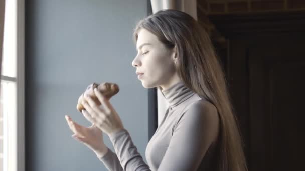 苗条优雅的年轻女子闻到并吃着甜可口的羊角面包的侧视图。早上站在窗边享用早餐的白种人美丽而自信的黑发女子的画像. — 图库视频影像