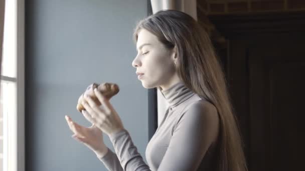 Widok z boku smukłej eleganckiej młodej kobiety pachnącej i jedzącej słodki, smaczny rogalik. Portret pewnej siebie pięknej brunetki rasy kaukaskiej cieszącej się śniadaniem rano stojąc przy oknie. — Wideo stockowe