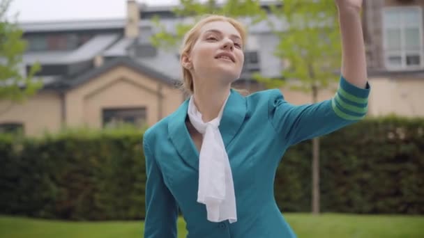 Retrato de encantadora mulher bonita magro em azul aeromoça uniforme acenando e sorrindo. Assistente de bordo caucasiano elegante confiante à espera de reunião ao ar livre. Turismo e viagens. — Vídeo de Stock