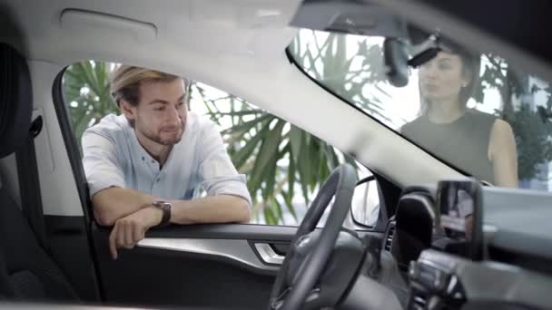 Retrato de hombre de negocios guapo sonriente mirando dentro de coche nuevo en la concesionaria como mujer hablando. Cliente caucásico admirando vehículo mientras concesionario explicar detalles. Comprador feliz en sala de exposición. — Vídeo de stock