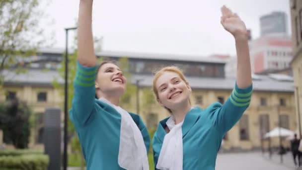 Positiva unga smala kvinnor i flygvärdinnas uniform vinkar och tittar upp. Porträtt av leende eleganta vita flygvärdinnor som står utomhus. Rese- och turismkoncept. — Stockvideo