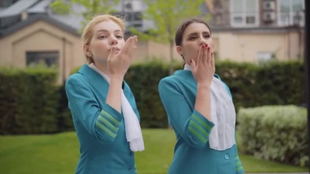 Unga eleganta vita flygvärdinnor som skickar luftkyssar och ler. Porträtt av två självsäkra vackra kvinnor i blå uniform stående på stadens gata. Begreppet turism och resor. — Stockvideo