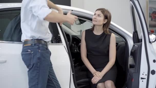 Mujer caucásica segura sentada en el asiento del pasajero en un coche nuevo y hablando con un distribuidor irreconocible en la sala de exposición. Retrato de una dama exitosa eligiendo el vehículo en el concesionario de automóviles. Negocios y riqueza. — Vídeos de Stock