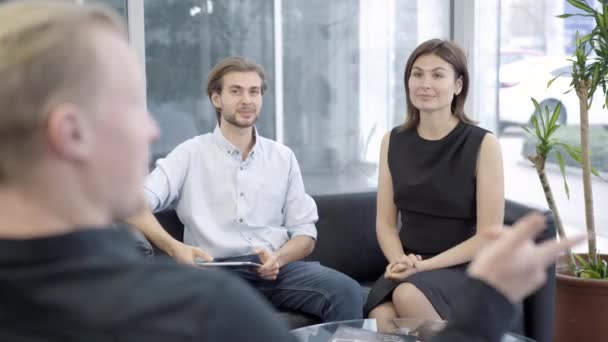 Hombre y mujer jóvenes exitosos sentados en el sofá y escuchando a un candidato caucásico irreconocible. Entrevista al potencial oficinista en interiores o reunión de socios comerciales. — Vídeos de Stock