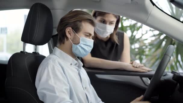 Νεαρός άνδρας με μάσκα προσώπου Covid-19 κάθεται στο κάθισμα του οδηγού στο αυτοκίνητο ως θολή γυναίκα μιλάει με τον αγοραστή στην αντιπροσωπεία. Πλάγια άποψη πορτρέτο του επιτυχημένου πελάτη που αγοράζει το όχημα κατά τη διάρκεια coronavirus. — Αρχείο Βίντεο