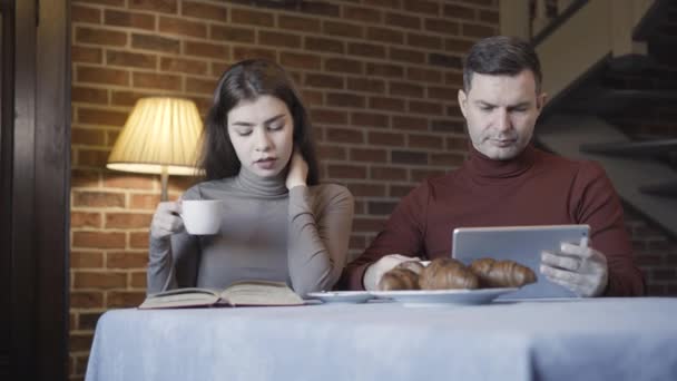 Όμορφος άνθρωπος surfing internet στο tablet ως γυναίκα ανάγνωση βιβλίο στο πρωινό στο σπίτι. Πλούσιο λευκό ζευγάρι πίνει καφέ το πρωί σε εσωτερικούς χώρους. Έννοια της οικογενειακής ζωής. — Αρχείο Βίντεο