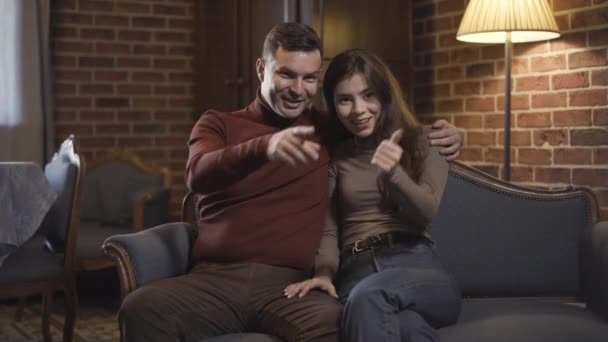 Позитивная обнимающая пара сидит в гостиной и смотрит телевизор. Счастливый богатый кавказский мужчина и молодая женщина наслаждаются отдыхом в выходные в помещении дома. Концепция роскоши и отдыха. — стоковое видео
