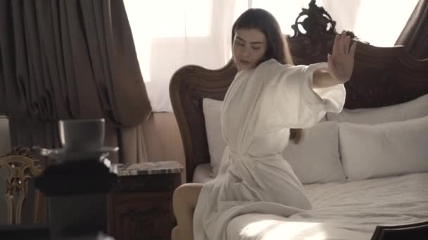 Vacker smal ung kvinna som sträcker sig på morgonen sittande på sängen. Sidovy porträtt av vacker brunett vit dam i vit badrock vakna upp i sovrum hemma eller på hotell. Kvinnlighet. — Stockvideo