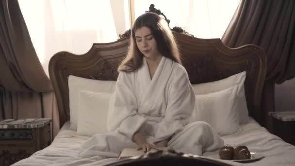 Mulher branca bonita magro sentado na cama com livro e comer croissant delicioso. Retrato de jovem senhora despreocupada desfrutando de manhã ensolarada no quarto em casa ou no hotel. — Vídeo de Stock