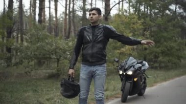 Genç yakışıklı Ortadoğulu motosikletçi arka planda kırık bir motosikletle yol kenarında otostop çekiyor. Bulutlu bir günde stresli bir adamın portresi. Yarış ve kırılma.