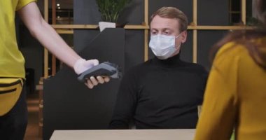 Covid-19 yüz maskeli genç beyaz adam restorandaki POS terminalinde akıllı telefon ödemesi yapıyor. Kendine güvenen koca, karısının koronavirüs salgınıyla olan yemeğini ödüyor. Sinema 4k ProRes Merkezi.