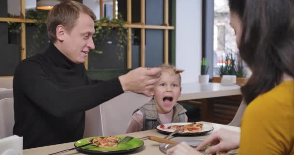 Glücklicher kaukasischer Vater, der seine Tochter im Restaurant mit einem Kochlöffel füttert, während die verschwommene Mutter vorne Pizza isst. Fröhlich entspannte Familie beim Abendessen in der Pizzeria. Cinema 4k ProRes Hauptquartier. — Stockvideo