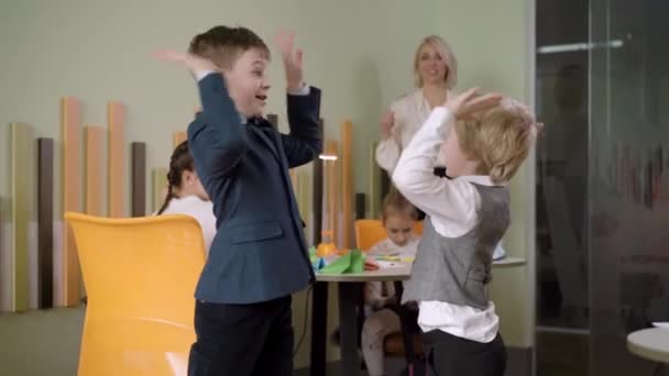Vreugdevolle blanke jongens geven high-five en lachen met wazige leraar en klasgenoten op de achtergrond. Positieve schooljongens die plezier hebben tijdens de les op de openbare school. Onderwijs en vrije tijd. — Stockvideo
