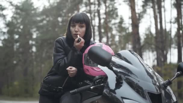 Porträtt av vacker säker motorcyklist tillämpa rött läppstift utomhus tittar i sidospegel. Brunett vit kvinna sitter på svart motorcykel och gör make-up. Skönhet och livsstil. — Stockvideo