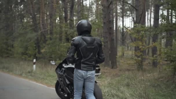 Młody pewny siebie motocyklista idzie do motocykla i siedzi na nim. Mężczyzna w skórzanej kurtce i butach, dżinsy i kask jazda na motocyklu na świeżym powietrzu w pochmurny jesień lub wiosenny dzień. Pojęcie hobby. — Wideo stockowe