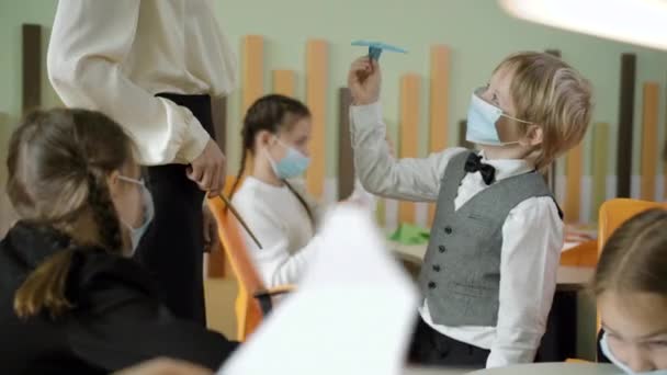 Lindo chico caucásico en máscara de la cara Covid hablando con el profesor delgado irreconocible y el lanzamiento de avión de papel. Lección de artes positivas en la escuela pública sobre la pandemia del coronavirus. Educación y estilo de vida. — Vídeos de Stock