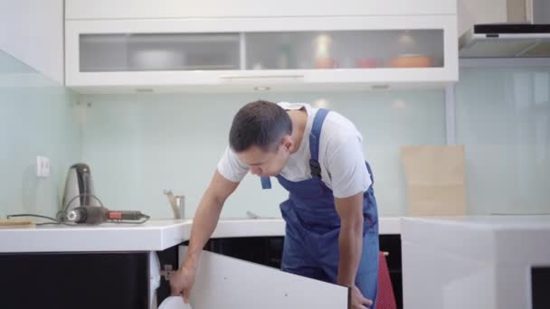 Jeune menuisier professionnel examinant la porte cassée de l'armoire de cuisine dans l'appartement. Brunette homme de service caucasien en uniforme bleu fixation rupture à plat. Concept d'occupation. — Video