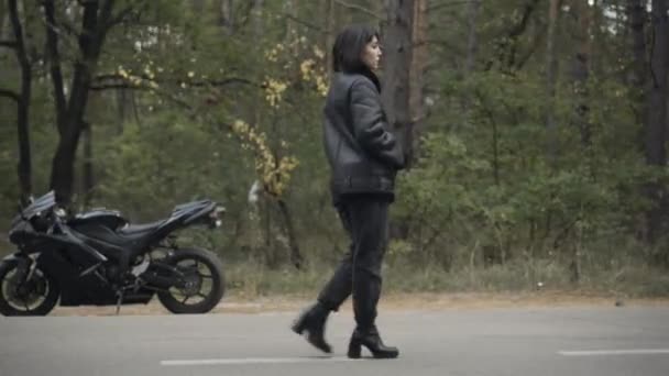 Bulutlu bir günde asfalt yolda yürüyen kendine güvenen bir kadının yan görüntüsü. Düşünceli beyaz motorsikletlinin bahar ya da sonbahar günlerinde açık havada dolaşırken geniş açılı görüntüsü. Yaşam tarzı ve yalnızlık. — Stok video