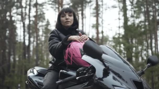 Bella giovane donna caucasica seduta su moto nera con casco rosa e guardando la fotocamera. Ritratto di pilota bruna fiducioso in posa all'aperto nella nuvolosa giornata primaverile o autunnale. Concetto di hobby. — Video Stock