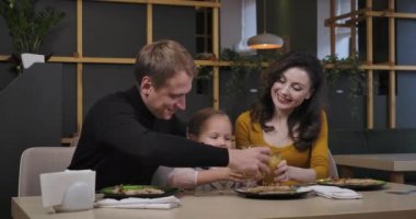 Olumlu baba, anne ve kız meyve suyu bardaklarını tokuşturup gülümseyerek başlarını çeviriyorlar. Mutlu Kafkasyalı erkek, kadın ve kız pizzacıda yemek yiyor. Sinema 4k ProRes Merkezi.