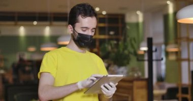 Tablete yazı yazan ve kameraya bakan Kafkas maskeli erkek garsonun portresi. Coronavirüs salgınında restoranda poz veren bir işçi. Yeni normal servis. Sinema 4k ProRes Merkezi.