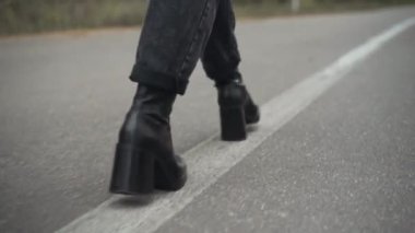 Kot pantolonlu kadın ayakları ve beyaz çizgi üzerinde yürüyen siyah deri topuklu ayakkabılar. Tanımlanamayan genç bir kadın bulutlu bir baharda ya da sonbahar gününde boş otoyolda geziniyor..