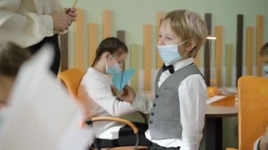 Sınıfta kağıt uçakla duran ve konuşan küçük çocuğun Covid maskesini ayarlayan tanınmamış bir öğretmen. Koronavirüs salgını sırasında ilkokul eğitimi. Tatlı çocuk ve bulanık sınıf arkadaşları..