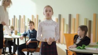 Kendine güvenen liseli kız kameraya bakıyor ve sınıfta şiir okuduğunu iddia ediyor. Arka planda bulanık sınıf arkadaşları ve öğretmeniyle devlet okulunda okuyan zeki beyaz kız portresi..