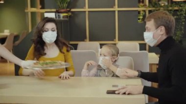 Covid maskeli pozitif aile restoranda tanınmayan bir garsonun yanında taze pişmiş lezzetli pizza getiriyor. Rahatlamış Kafkasyalı erkek, kadın ve kız Coronavirus salgınında pizzacıda yemek yiyorlar..