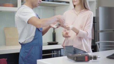 Marangoza ya da elektrikçiye hizmet için ödeme yapan tanınmamış bir kadın. Gülümseyen beyaz bir bayan, apartmanın mutfağında duran profesyonel servis elemanına para veriyor. Ev onarımı.